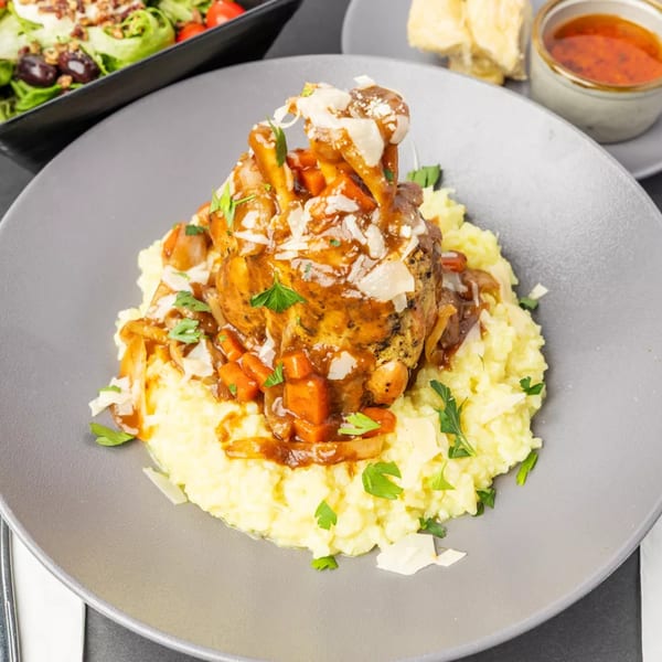 Braised lamb shank with vegetables and parmesan cheese over creamy risotto, with a salad in the background.