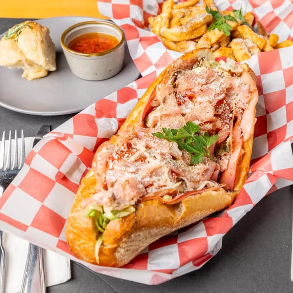 A large Italian sub sandwich, fries, and a side of dipping sauce on a checkered paper.