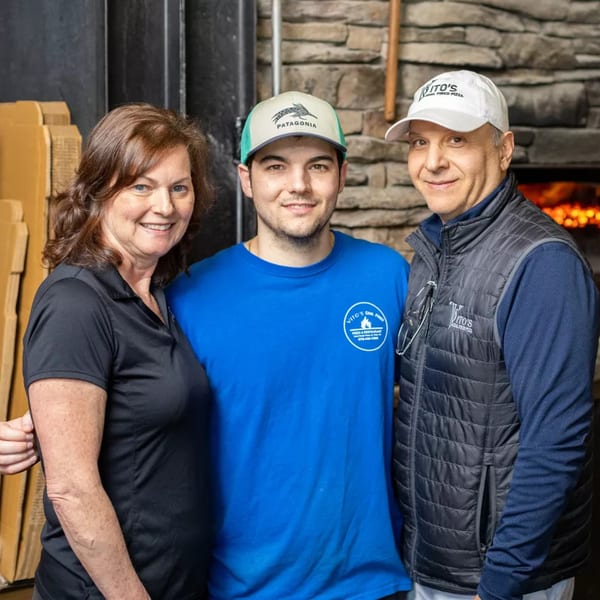 Three smiling people, likely owners or staff, stand in front of Vito's coal-fired pizza oven.