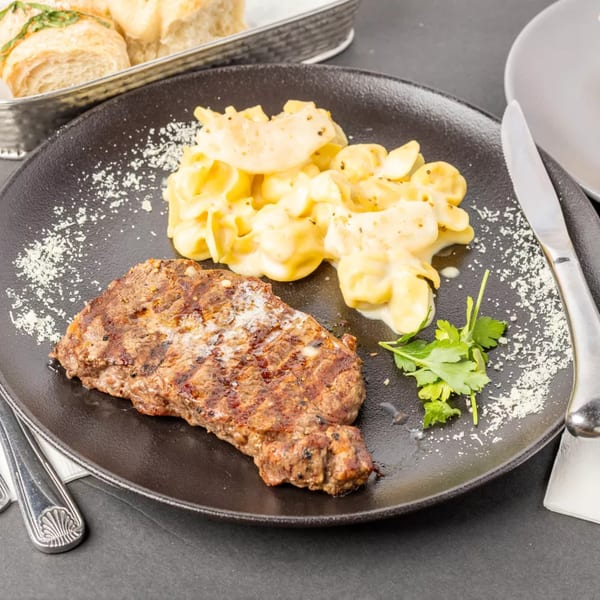 Grilled steak and creamy macaroni and cheese on a black plate, garnished with parsley and parmesan.